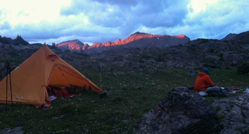a person sits outside an orange tent while the sun rises, shining light on the mountains in the background